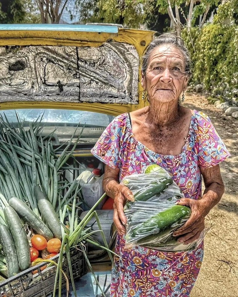 A woman from the community holding a bag of fresh zucchini beside a car full of produce
